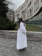 Woman in a long white faux fur coat standing by a modern building.