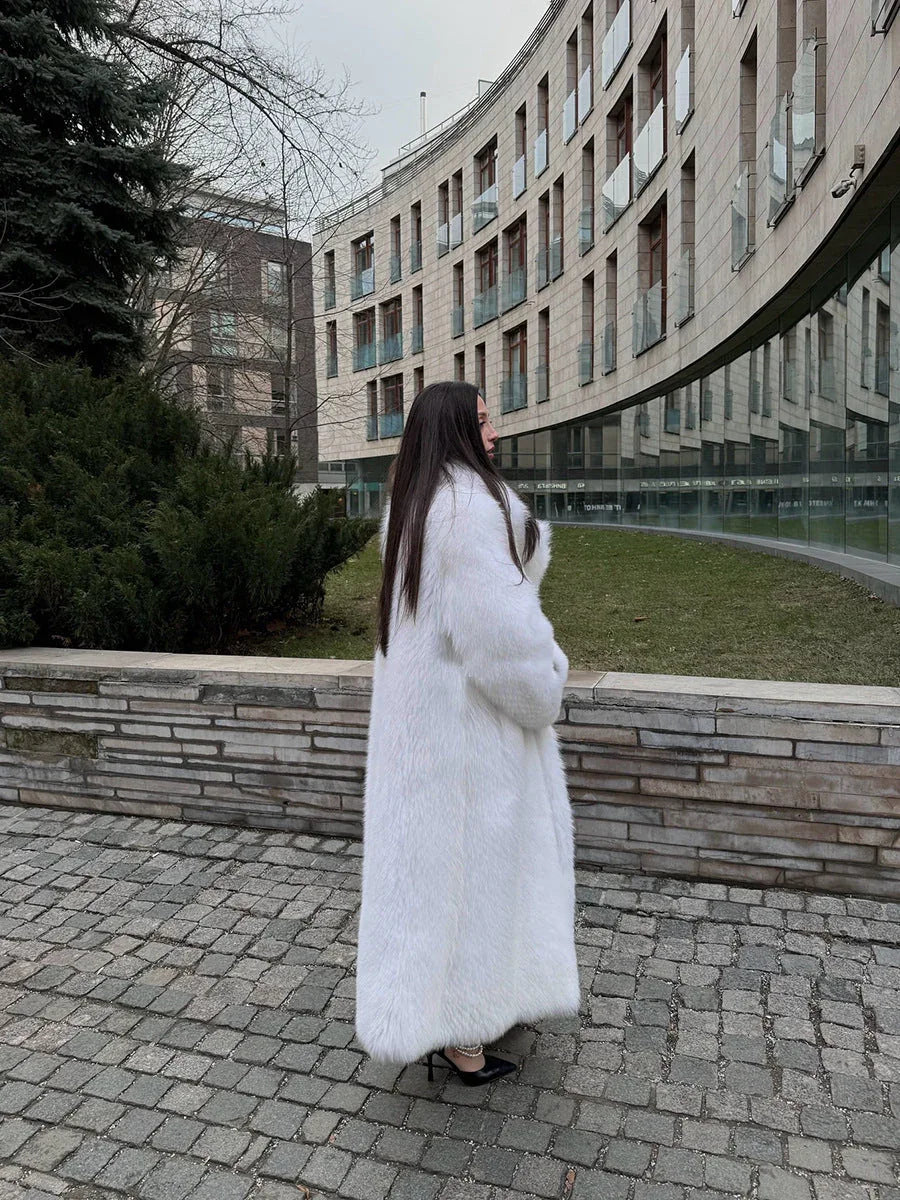 Woman in a long white faux fur coat standing by a modern building.