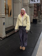 Woman wearing a cream faux fur cropped jacket with a lapel collar and long sleeves, paired with grey pleated pants and a white knit beanie.