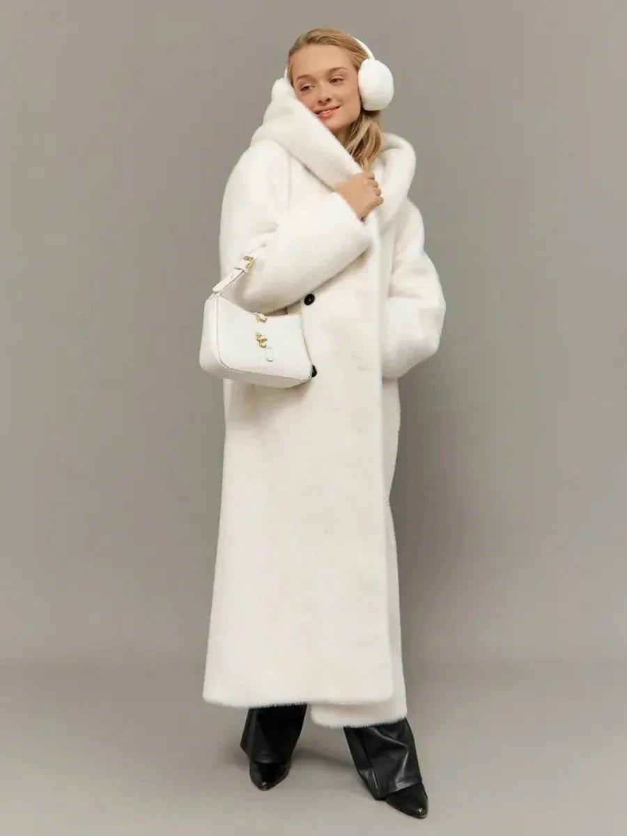 Woman wearing a long white hooded faux fur coat with a belted waist, black trousers, and white earmuffs. Main image