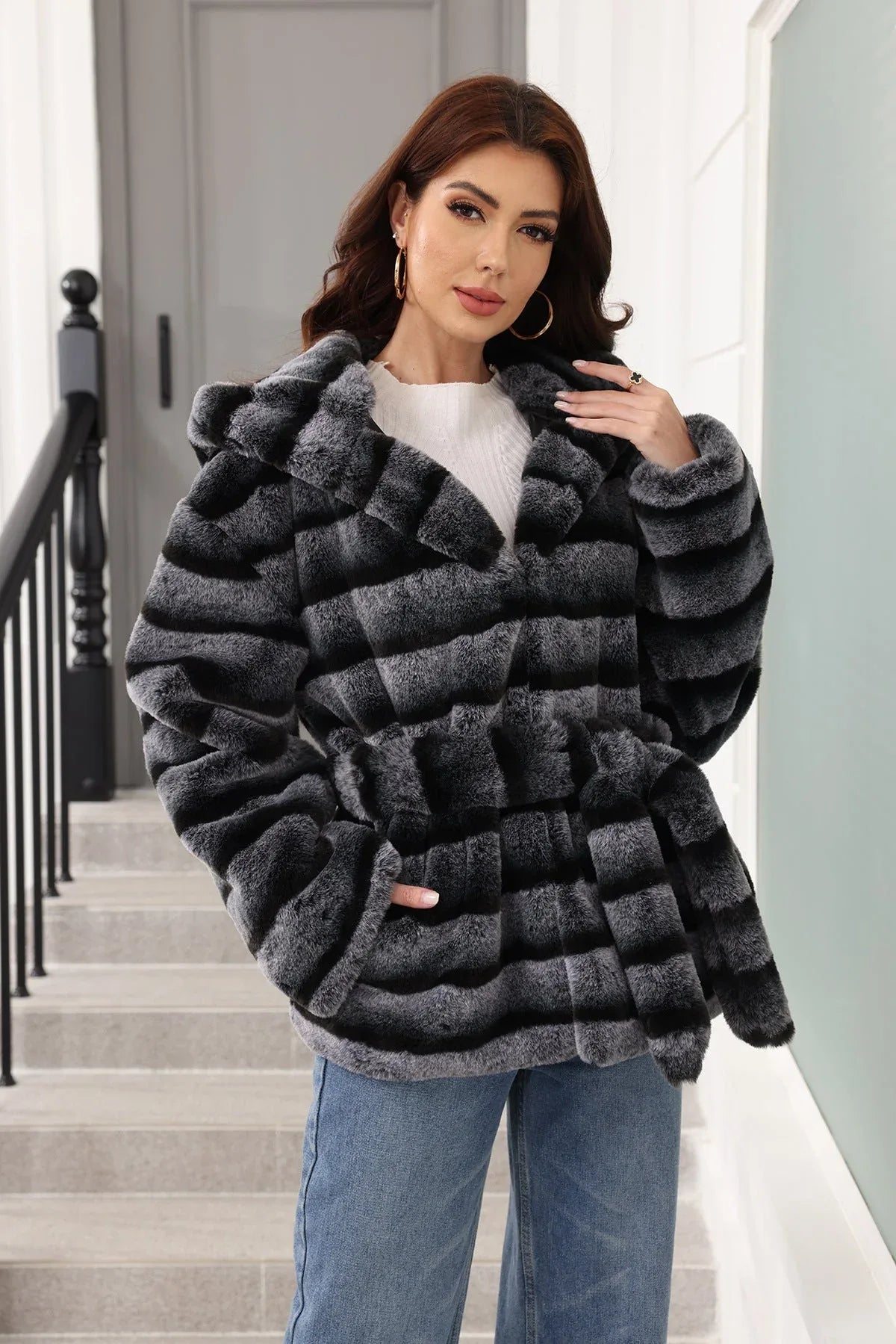 Woman posing in a hooded, belted, grey and black striped faux fur jacket, touching her hair.