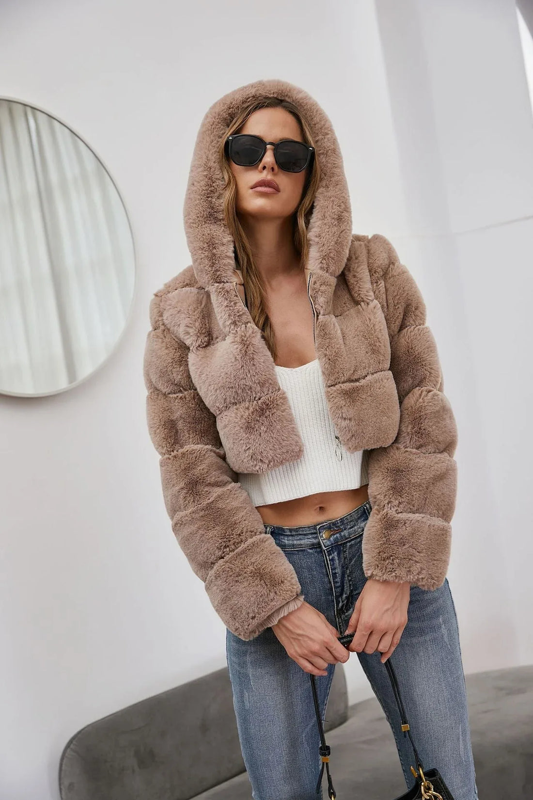 Woman wearing a cropped taupe faux fur hooded jacket with a white crop top and blue jeans.