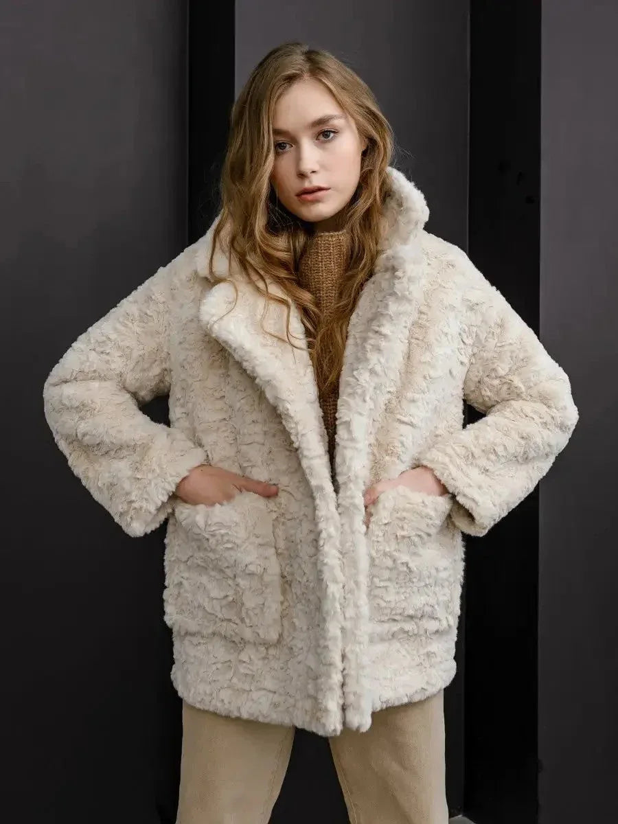 Model wearing a cream faux fur coat with notch lapels and patch pockets, paired with tan trousers and a brown knit sweater.