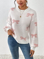 Front view of a woman in a white knit sweater with a pink bow print and crew neckline, holding her hand on her hip.