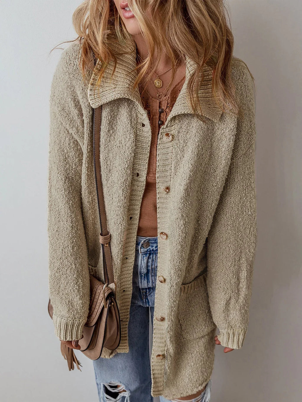 Woman in a beige textured knit cardigan, brown top, and blue jeans, accessorized with a crossbody bag.