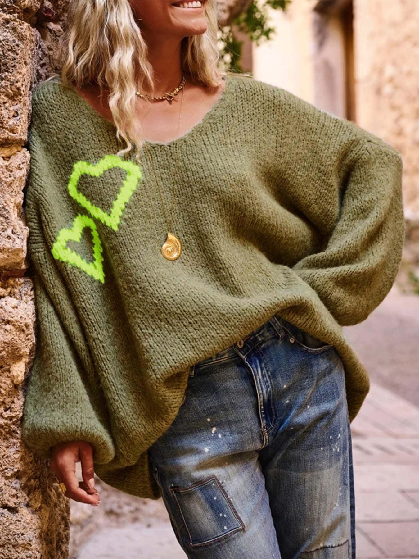Woman wearing an oversized olive green V-neck knit sweater with embroidered neon yellow heart graphics on the left chest, paired with distressed denim jeans.