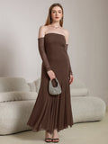 Woman holding a small embellished handbag while wearing a long brown off-the-shoulder dress with sheer sleeves