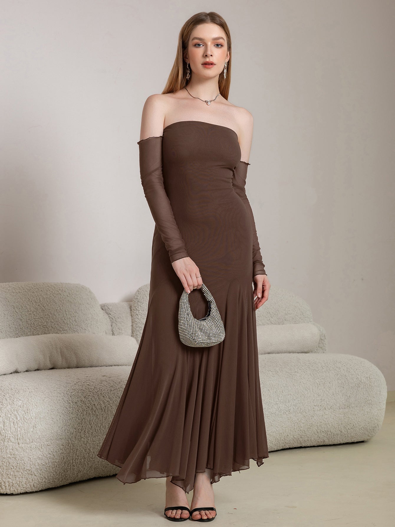 Woman holding a small embellished handbag while wearing a long brown off-the-shoulder dress with sheer sleeves