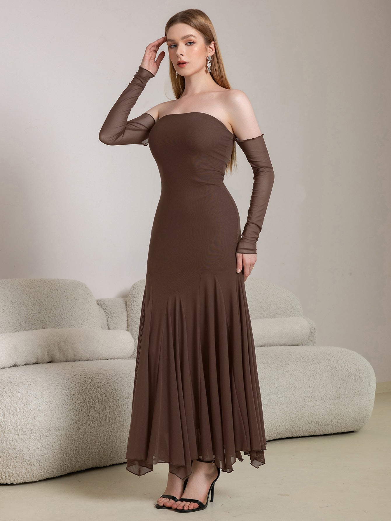 Woman adjusting her hair while wearing a long brown off-the-shoulder dress with sheer sleeves