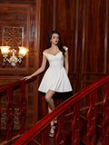 Woman in a white off-the-shoulder dress with a fitted bodice and pleated skirt, leaning on a wooden railing.