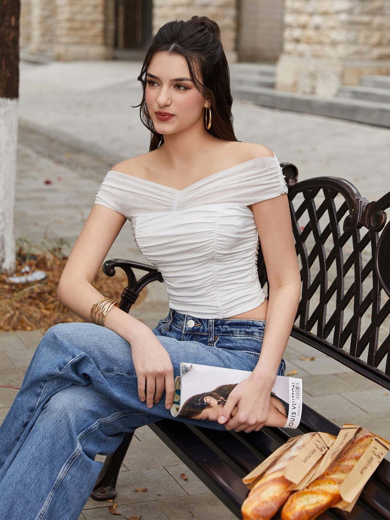 Woman holding sunglasses and a magazine while wearing a white off-the-shoulder ruched crop top and blue jeans