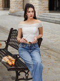 Woman sitting on a bench wearing a white off-the-shoulder ruched crop top and blue jeans