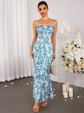 Woman wearing a long, strapless, white dress with a blue floral print, featuring a sweetheart neckline and a mermaid silhouette. She is holding a small woven bag.