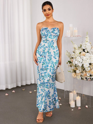 Woman wearing a long, strapless, white dress with a blue floral print, featuring a sweetheart neckline and a mermaid silhouette. She is holding a small woven bag.