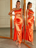 Model wearing an orange satin one-shoulder maxi dress with cut-out details and a high slit, standing with her hand on her hip.