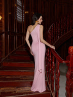 Back view of a woman in a pink one-shoulder maxi dress with draped detailing on a wooden staircase.
