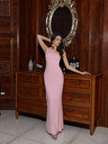 Woman in a pink one-shoulder maxi dress with draped detailing posing next to a wooden dresser.