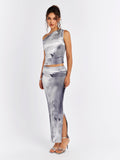 Side view of a woman wearing a grey and white abstract print asymmetrical sleeveless top and matching maxi skirt with a high slit.