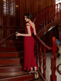 Back view of a woman in a red one-shoulder mermaid gown holding a wine glass on a wooden staircase.