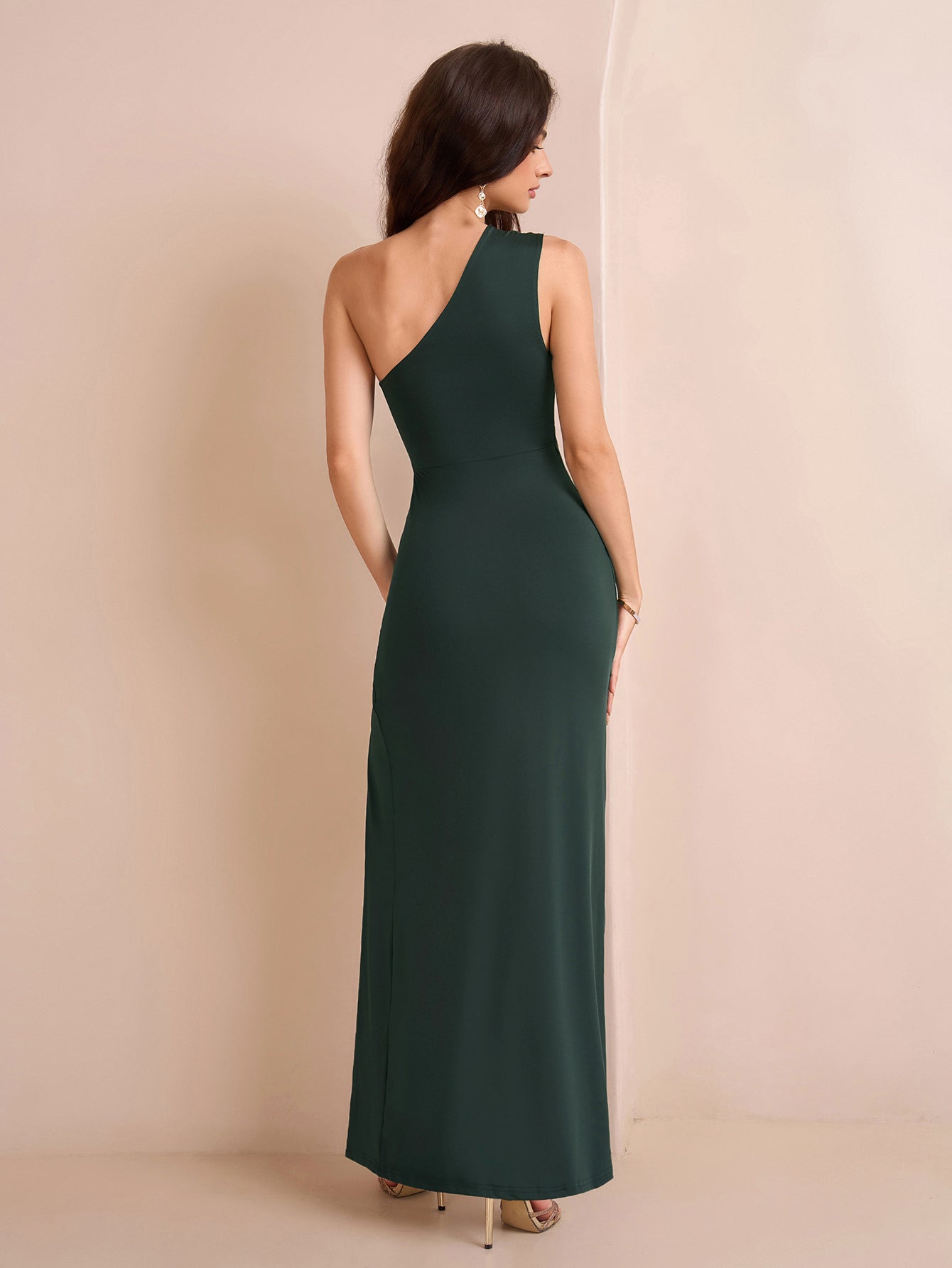 Back view of a woman in a dark green one-shoulder jersey knit column maxi dress with a high slit