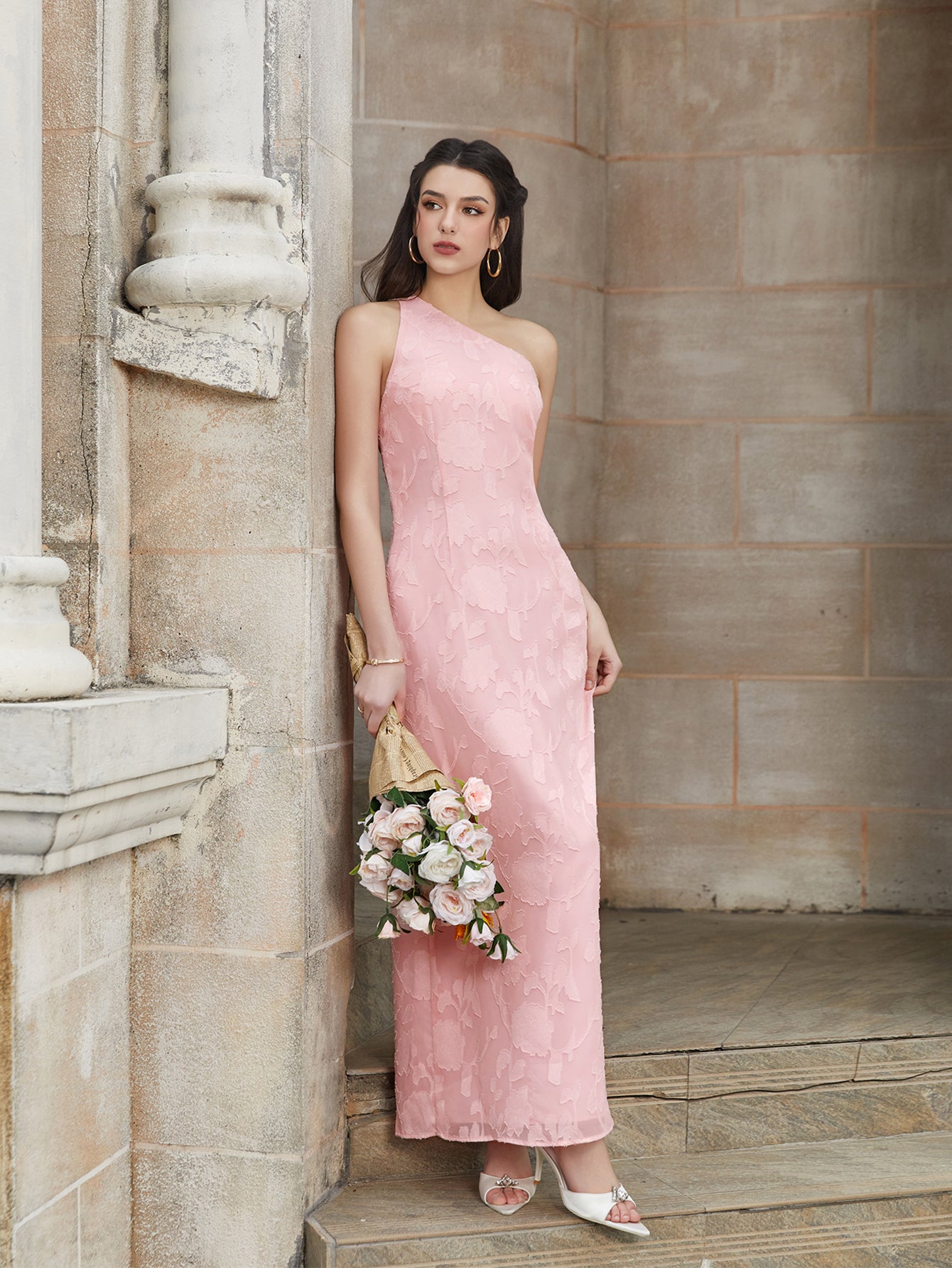 A woman poses in a pink one-shoulder floral burnout maxi dress, leaning against a stone wall.