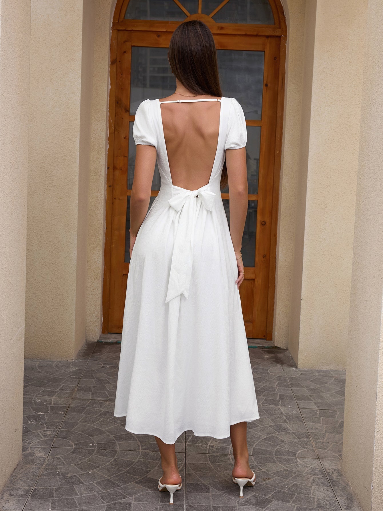 Back view of a woman in a white midi dress with an open back tied with a bow.