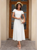 Front view of a woman in a white midi dress and straw hat, holding a beige handbag.