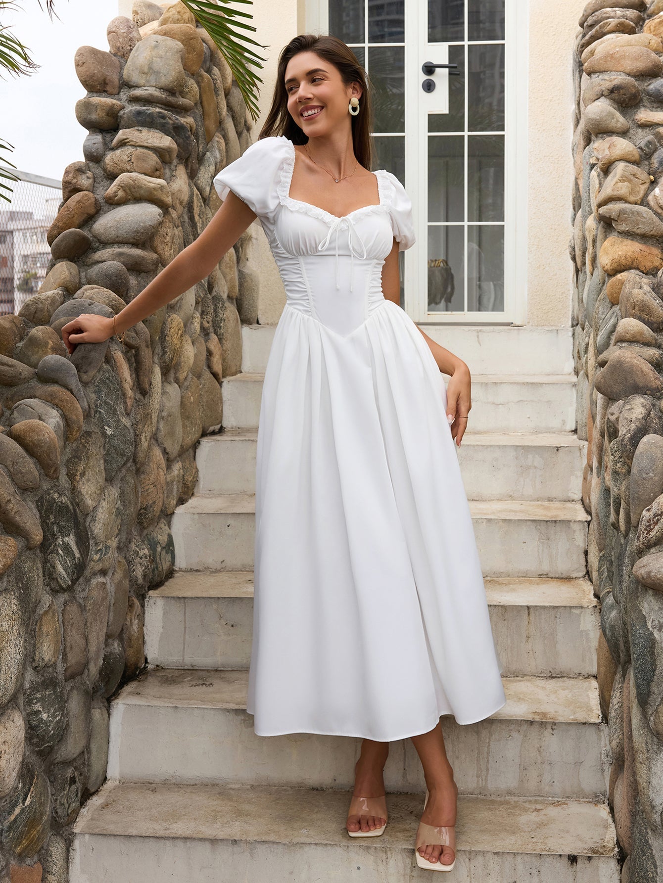 Woman smiling in a white puff sleeve midi dress with a square neckline and corset bodice, on outdoor stairs.