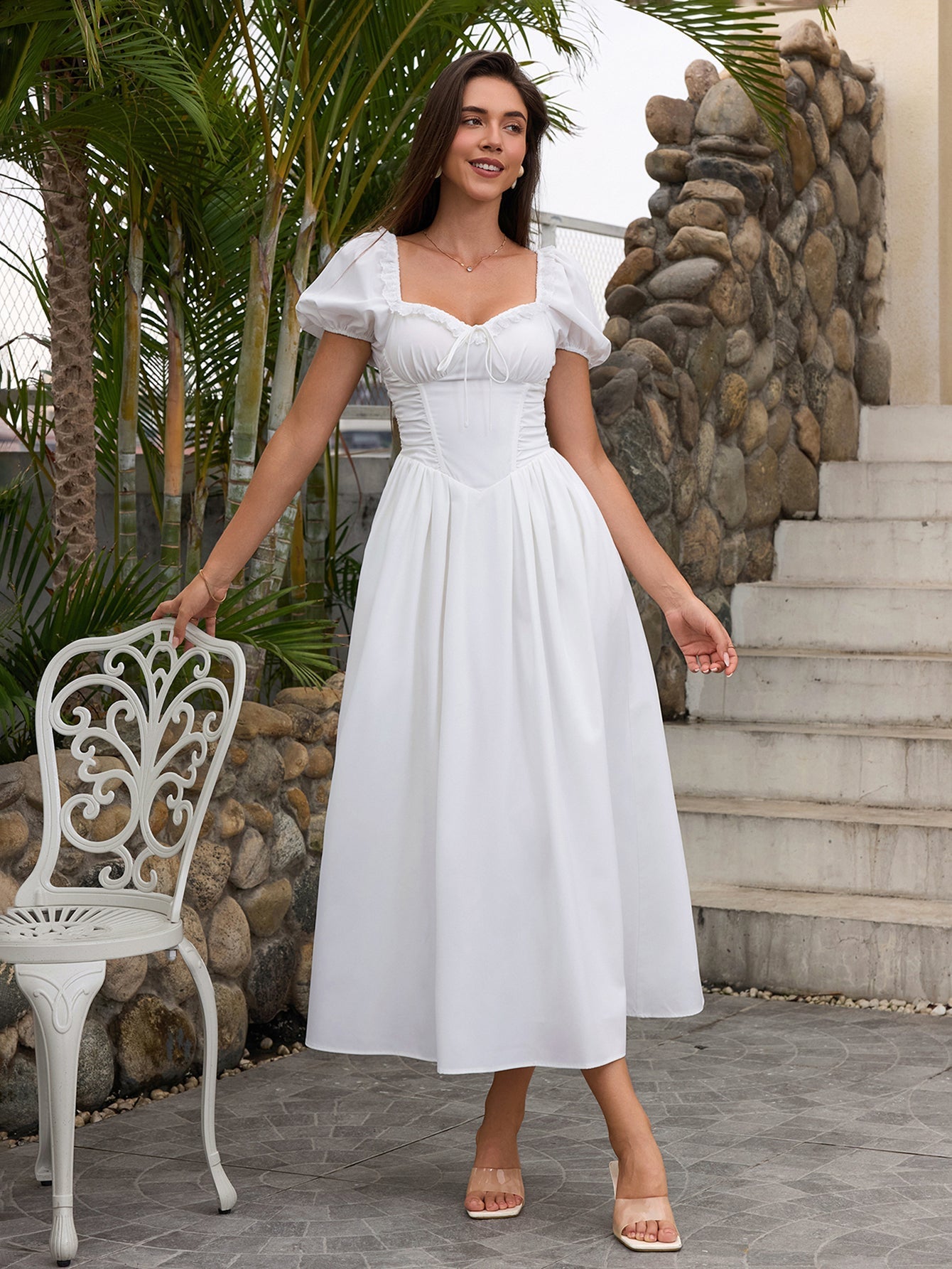 Woman in a white puff sleeve midi dress with a square neckline and corset bodice, standing next to a chair outdoors.