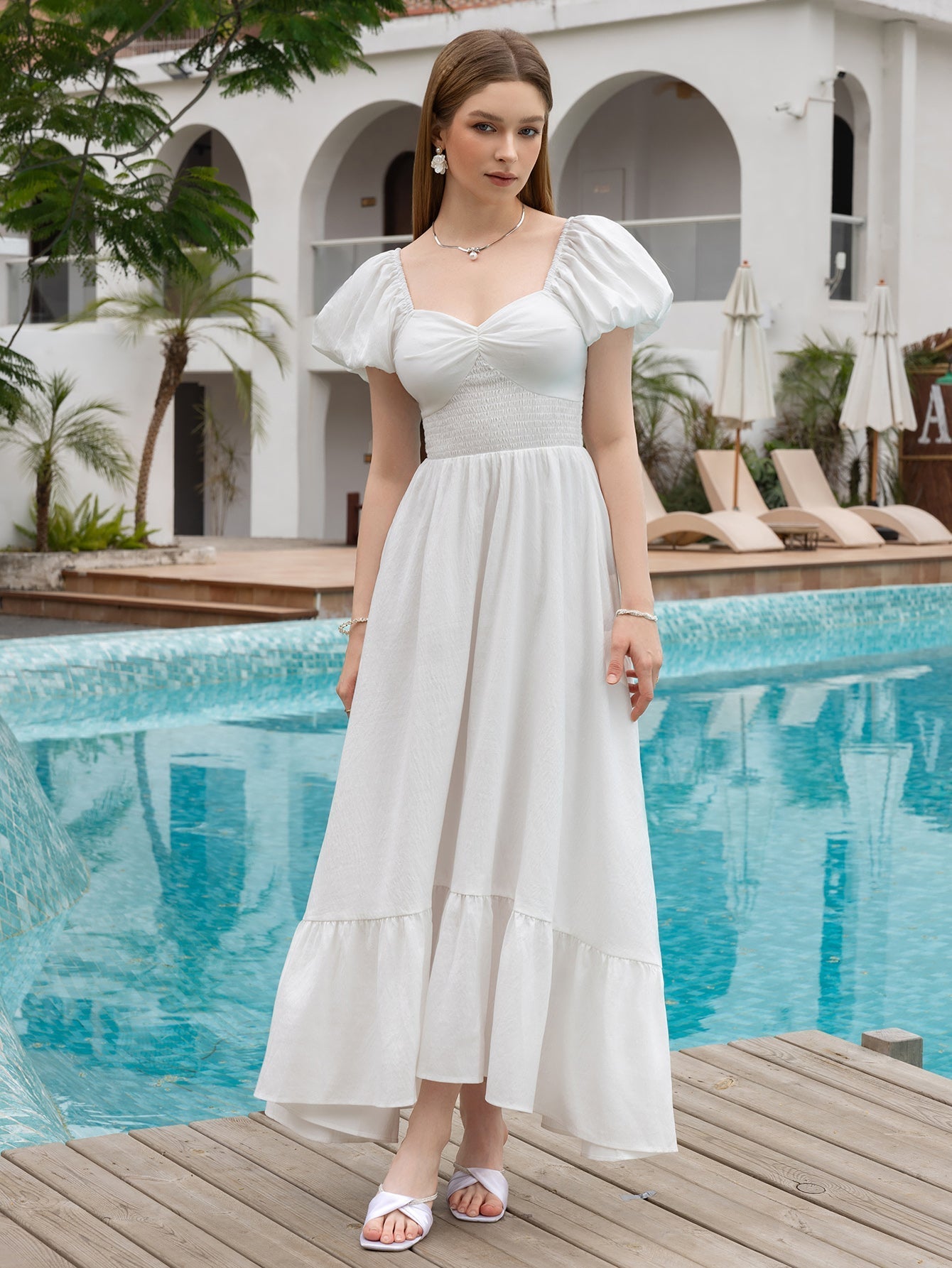 Woman wearing a white puff sleeve maxi dress with a smocked waist standing by a pool.
