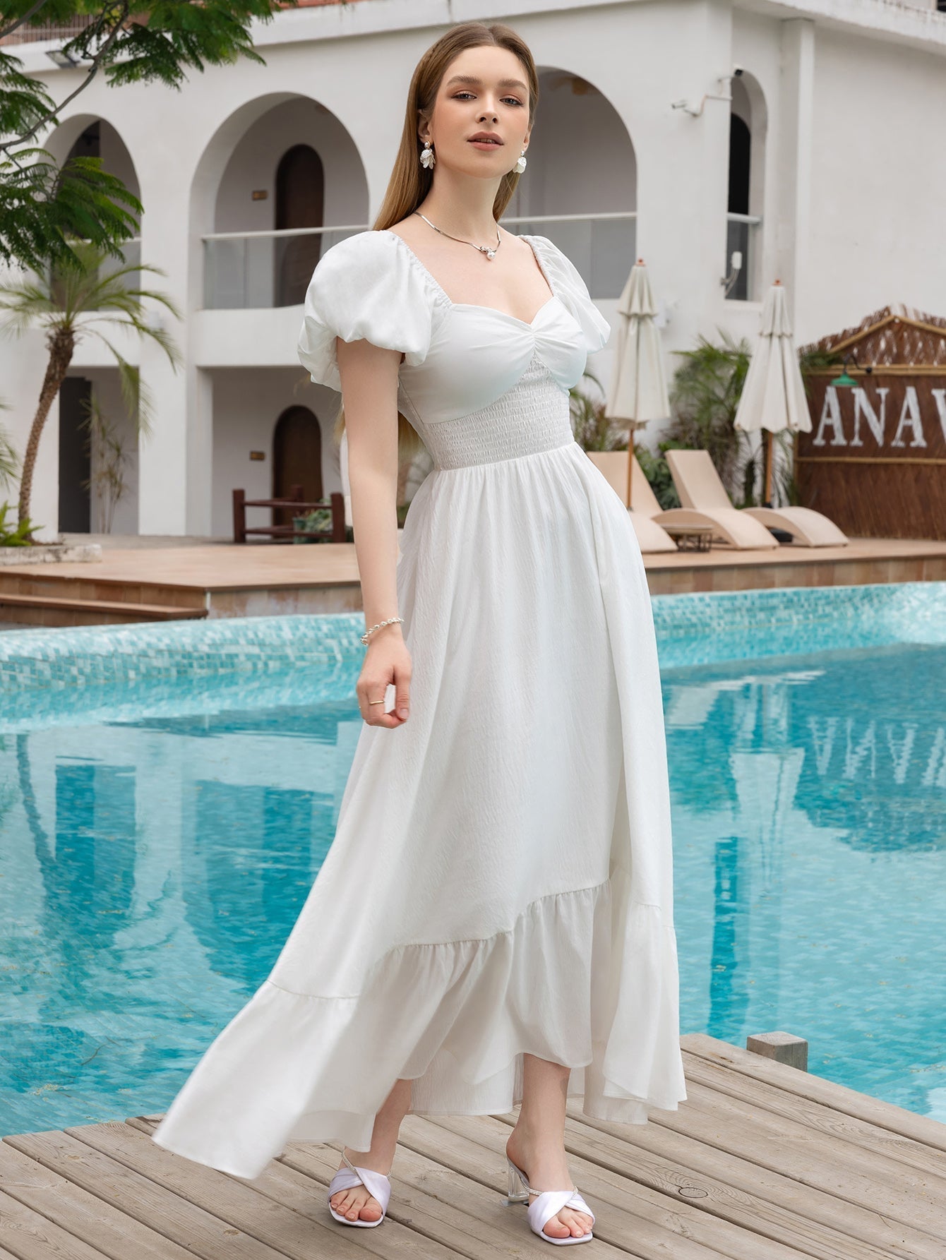Woman in a white puff sleeve maxi dress with a sweetheart neckline and tiered hem posing by a pool.