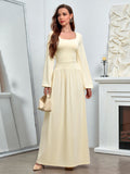 Woman in a pale yellow maxi dress with a square neckline and long sleeves, holding a handbag.