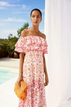 Woman wearing a white and pink off-the-shoulder midi dress with ruffle trim and embroidered circular patterns, holding an orange mesh clutch.