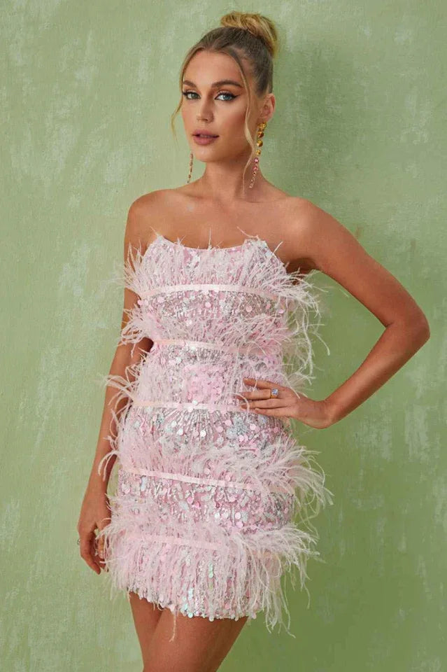 Woman in a pink strapless sequin mini dress with feather trim and ribbon details, posing with hand on hip. Main image