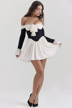 Woman posing in a black and cream off-the-shoulder dress featuring a bow detail and pleated skirt.