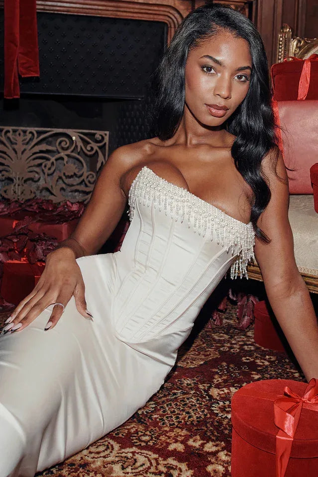 Close-up of a woman wearing a white satin strapless corset midi dress with beaded trim and a slit hem, seated among red gift boxes.