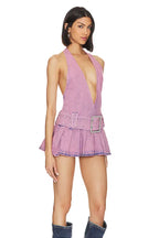 Side view of a woman wearing a lavender washed denim halter neck mini dress with a plunging V-neckline, a wide embellished belt, and a pleated skirt, paired with denim boots.