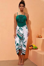 Woman wearing a green one-piece swimsuit with a large bow detail at the bust and a white and green leaf-print sarong wrap skirt.