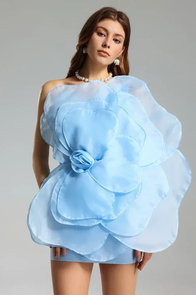 Woman wearing a light blue one-shoulder mini dress with a large organza floral applique on the front. Main image