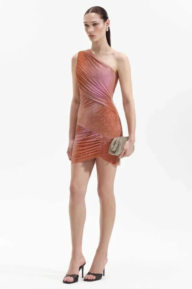 Woman in a shimmering one-shoulder mini dress with ruching and asymmetrical hem, holding a metallic clutch and wearing heeled mules.