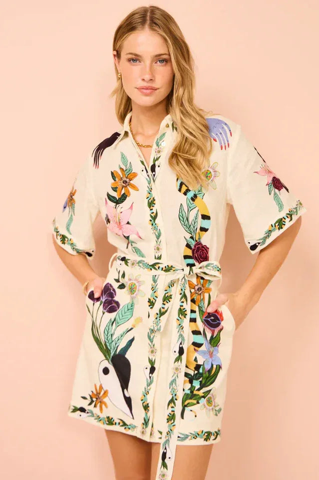Woman wearing a cream-colored short-sleeved shirt dress with a colorful floral and animal print, belted at the waist. Main image