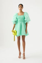 Woman wearing a mint green organza mini dress with puffy sleeves and a square neckline, holding a green clutch