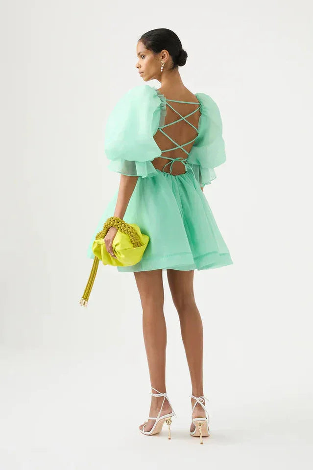 Back view of a woman wearing a mint green organza mini dress with puffy sleeves and an open back with criss-cross ties