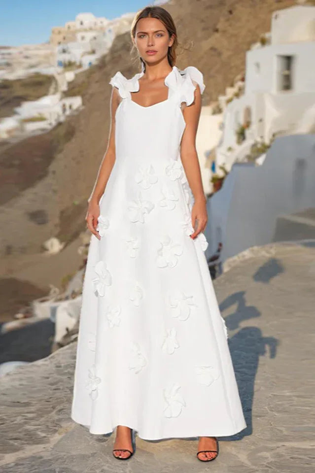 Woman wearing a white floor-length A-line dress with 3D floral appliqués and ruffled shoulder straps, standing outdoors with white buildings in the background.