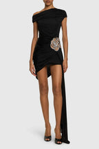 Woman wearing a black one-shoulder asymmetrical mini dress with draped fabric and a floral embellishment on the hip, featuring a high slit and a long flowing train.
