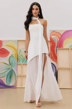 Woman wearing a white halter neck maxi dress with a high slit and sheer overlay, standing in front of colorful abstract paintings.