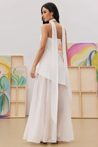 Back view of a woman in a white halter neck maxi dress with a draped scarf detail and sheer overlay.