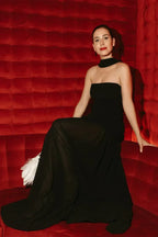 Woman wearing a black strapless gown with a sheer skirt, sitting on a red velvet couch.