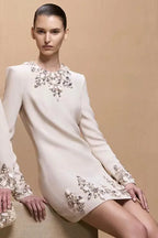 Woman wearing a cream long-sleeve mini dress with ornate crystal and pearl embellishments around the neckline, cuffs, and hem.