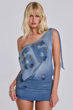 Model wearing a light blue distressed denim mini dress with stud embellishments and a single shoulder strap with tie detail.